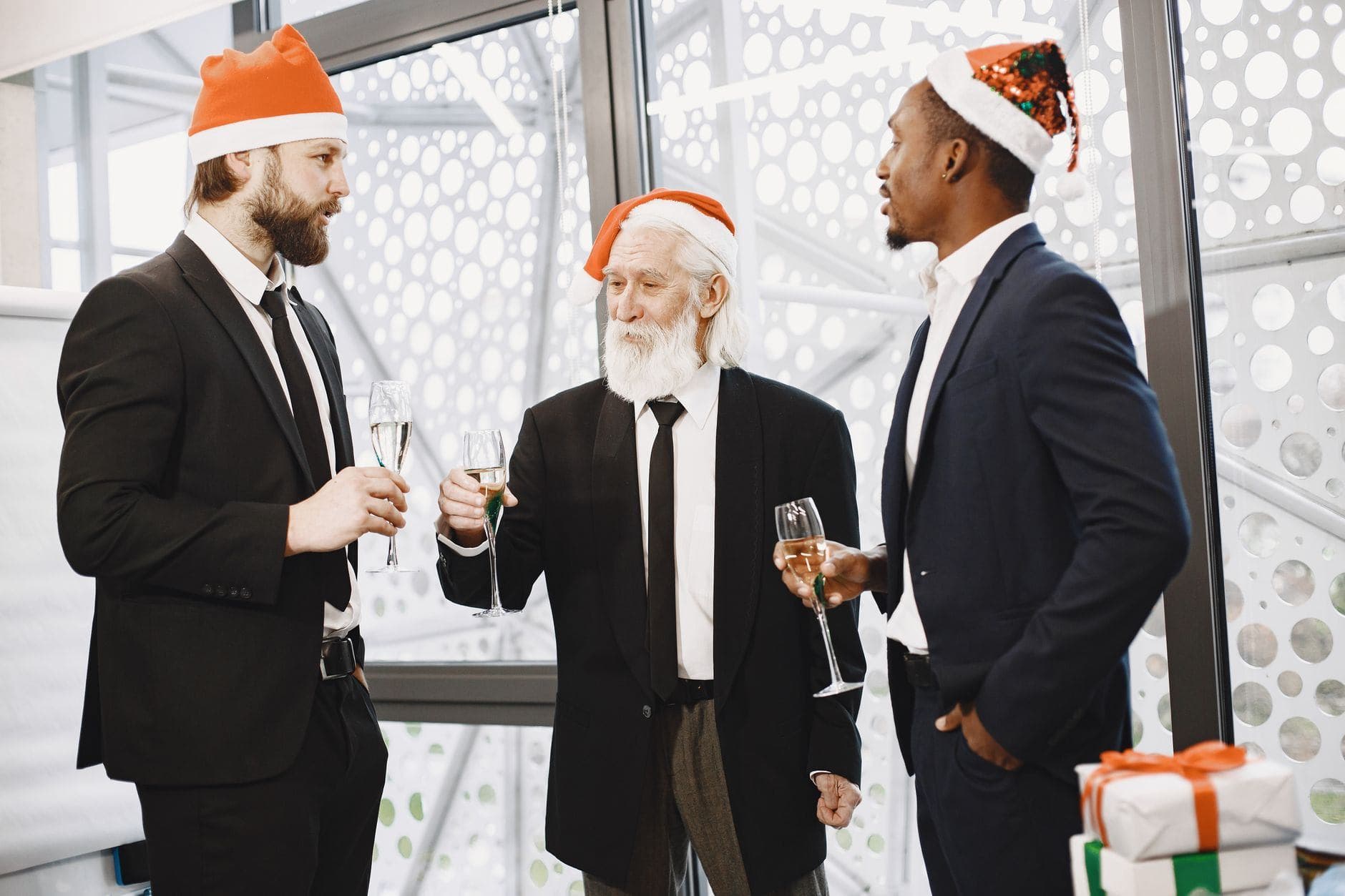 Three businessmen in suits celebrate Christmas with champagne indoors, wearing festive hats.
